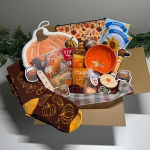 May include: A cardboard gift box containing a pumpkin-shaped potholder, brown socks with yellow pumpkin patterns, a zippered pouch with leaf prints, Stash tea packets, a small orange bowl, a candle with a wooden lid, and various seasonal stickers.