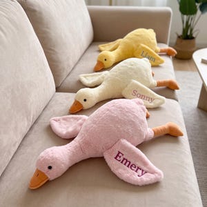 May include: Three plush goose toys in pink, cream, and yellow, each featuring an orange beak and embroidered names on the wing. The pink goose is labeled Emery, the cream goose is labeled Sonny, and the yellow goose is labeled Liam.