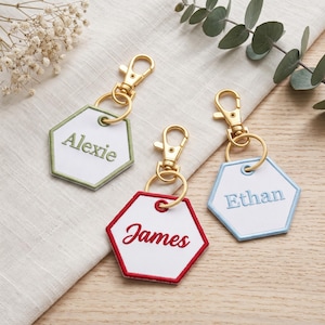 May include: Three hexagonal keychains with gold-tone lobster clasps. Each white fabric tag features a colored border and embroidered name: Alexie in green, James in red, and Ethan in light blue. The keychains are arranged on a light surface.
