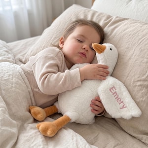 May include: A plush white goose toy with orange feet and an orange beak. The name Emery is embroidered in pink thread on one of the goose's wings. The toy has a soft, fuzzy texture and is designed with a long neck and outstretched wings.