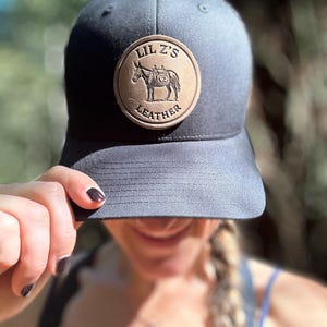 May include: A dark gray baseball cap with a circular leather patch. The patch features a detailed illustration of a donkey carrying a pack, with the text "LIL Z'S LEATHER" around the edge. The cap has a curved brim and a structured crown.