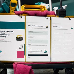 May include: A yellow janitorial cart holding three white clipboards with printed operational documents. The documents feature text including Clean Standard Crew Operations Library, Make a reliable clean teachable, and Operations setup, along with small icons of cleaning tools and a pink cloth draped over the cart.