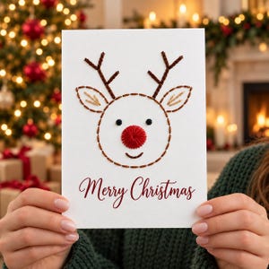 May include: A white greeting card featuring a reindeer face created with brown and tan embroidery thread. The reindeer has a prominent red stitched nose, black eyes, and the text Merry Christmas printed in a dark red cursive font below the face.