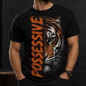 Könnte beinhalten: Ein schwarzes Kurzarm-T-Shirt mit einem Grafikdruck eines Tigergesichts auf der rechten Seite und dem Wort POSSESSIVE, das in großen, abgenutzten orangefarbenen Großbuchstaben vertikal auf der linken Seite des Brustbereichs steht.