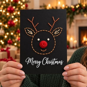 Puede incluir: Una tarjeta de felicitación negra con una cara de reno creada con bordados en marrón y beige. El reno tiene una prominente nariz roja bordada, dos pequeños acentos circulares blancos para los ojos y el texto Merry Christmas en blanco.