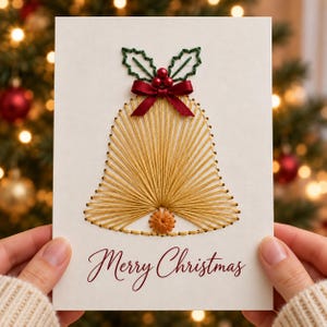 May include: A white greeting card featuring a gold string art bell design. The bell is accented with a dark red satin ribbon bow, three small red beads, and green embroidered holly leaves. The text Merry Christmas is printed in red script below.