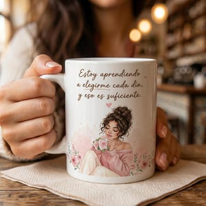May include: White ceramic mug featuring a watercolor illustration of a woman holding flowers, with a pink and floral design. The mug has the text "Estoy aprendiendo a elegirme cada día, y eso es suficiente."