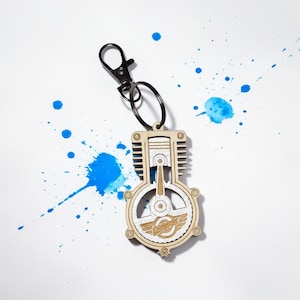 May include: A wooden keychain in the shape of an engine, with a silver-colored metal ring and clasp. The engine design features a piston and the word "Garage" with wings. The background has blue paint splatters.