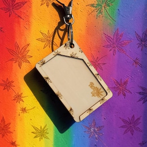 May include: Wooden keychain with a rectangular frame and a metal clasp. The frame is decorated with cannabis leaf designs. The background is a rainbow gradient with cannabis leaf patterns. The text "Roll Up" is visible on the keychain.