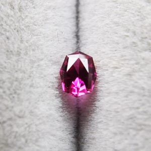以下が含まれることがあります： 鮮やかな深いマゼンタピンク色の、盾の形をしたファセットカットの宝石。石は対称的なカットが施されており、複数の三角形や台形のファセットが光を反射して、表面に輝きを生み出しています。