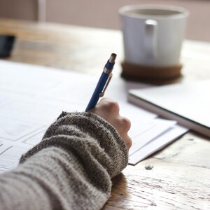 May include: A blue pen poised above white paper on a wooden table. A white mug and a coaster are in the background, along with a spiral-bound notebook. The image depicts a scene of writing or drawing.