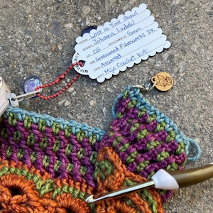 以下が含まれることがあります： オレンジ、緑、紫の糸を使ったショールを編んでいる様子をクローズアップ。かぎ針が使われています。「Lost in Time Shawl」と「Johanna Lindahl」と書かれたタグがあります。