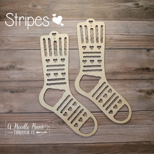 Puede incluir: Dos telares de madera para tejer calcetines con un patrón de rayas y corazones. Los telares están sobre un fondo de madera. El texto "Stripes" está en la parte superior de la imagen.