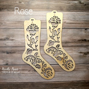 Puede incluir: Dos formas de bloqueo de calcetines de madera con un diseño de rosa. Las formas están etiquetadas con "M" para tamaño mediano. El texto "Rose" está en la parte superior de la imagen. El texto "A Needle Runs Through It" está en la parte inferior de la imagen.