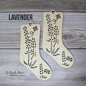 May include: Two wooden ornaments shaped like stockings with a cut-out design of lavender flowers, bees, and butterflies. The ornaments are labelled "LAVENDER".