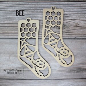 May include: Two wooden sock ornaments with a bee and honeycomb design. The text "BEE" is at the top of the image.