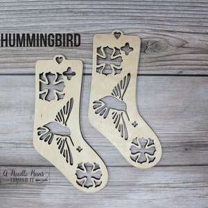 May include: Two wooden Christmas stocking ornaments featuring a hummingbird design and snowflake accents. The ornaments are unfinished and ready to be painted or stained. The text "HUMMINGBIRD" is printed at the top of the image.