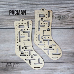 May include: Two wooden sock knitting looms shaped like Pac-Man characters. The looms are made of light brown wood and have holes for knitting needles. The text "PACMAN" is printed at the top of the image.