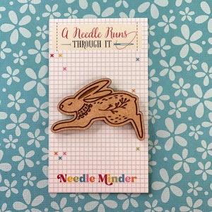 Puede incluir: Mantenga agujas de madera con un conejo marrón con detalles florales. El texto "A Needle Runs Through It" y "Needle Minder" está impreso en la tarjeta.