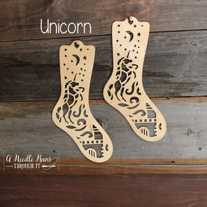 May include: Two wooden sock knitting looms featuring a unicorn, moon and stars design. The text "Unicorn" is above the looms. The text "A Needle Runs Through It" is below the looms.
