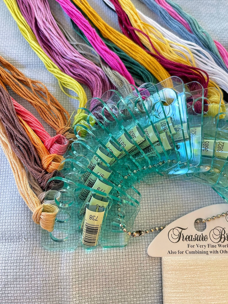 May include: A close-up of a set of plastic thread organizers with a variety of colored embroidery floss spools. The organizers are labeled with numbers, and the floss is arranged in a rainbow pattern. The text on the label reads "Treasure Box For Very Fine Work Also for Combining with Other Threads".