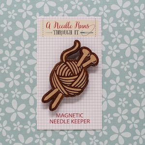 Könnte beinhalten: Magnetischer Nadelhalter aus Holz in Form einer Wollknäuel mit zwei herausstehenden Stricknadeln. Der Text "A Needle Runs Through It" ist auf der Karte hinter dem Nadelhalter gedruckt. Der Text "Magnetic Needle Keeper" ist unter dem Nadelhalter gedruckt.