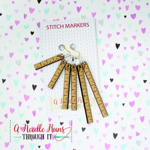 Pode incluir: Conjunto de seis marcadores de ponto de madeira com uma r&eacute;gua de cada lado. Os marcadores s&atilde;o presos a um anel dourado. A imagem tem um fundo de cora&ccedil;&otilde;es rosa, azuis e cinzentos. O texto "Stitch Markers" est&aacute; no topo da imagem. O texto "A Needle Runs Through It" est&aacute; na parte inferior da imagem.