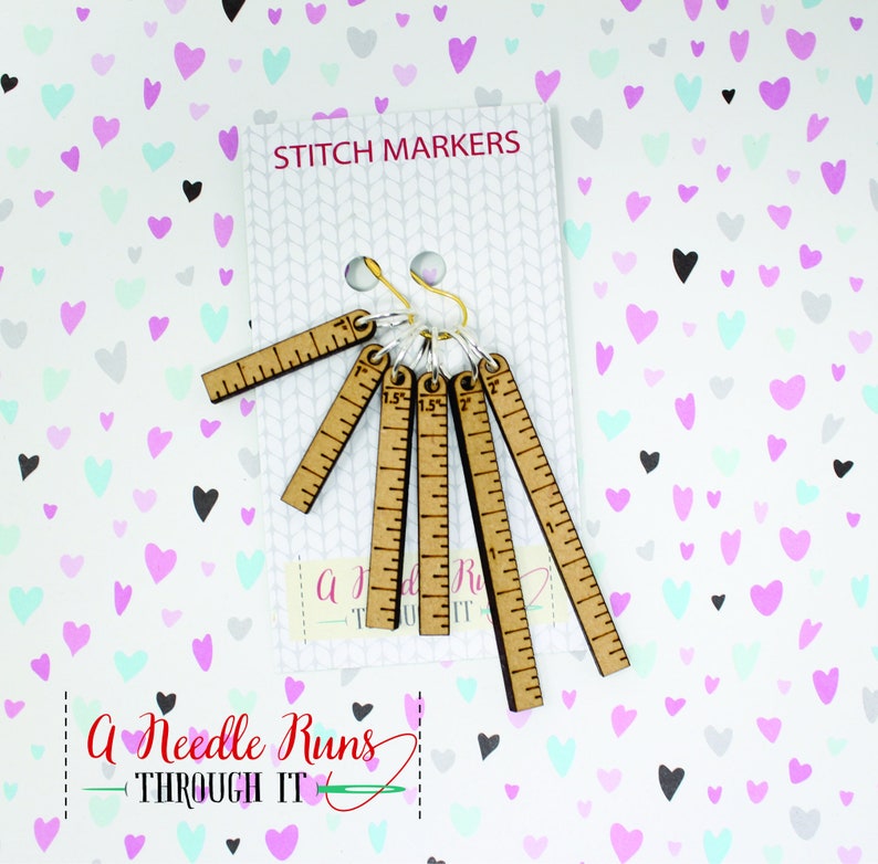 Pode incluir: Conjunto de seis marcadores de ponto de madeira com uma r&eacute;gua de cada lado. Os marcadores s&atilde;o presos a um anel dourado. A imagem tem um fundo de cora&ccedil;&otilde;es rosa, azuis e cinzentos. O texto "Stitch Markers" est&aacute; no topo da imagem. O texto "A Needle Runs Through It" est&aacute; na parte inferior da imagem.