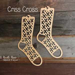Könnte beinhalten: Zwei sockengestaltige Stricknadelhalter aus Holz mit einem Kreuzmuster. Der Text "Criss Cross" befindet sich über den Haltern. Der Text "A Needle Runs Through It" befindet sich unter den Haltern.