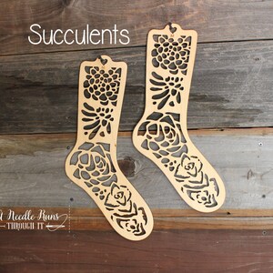 May include: Two wooden sock blockers featuring a succulent design. The blockers are made from natural wood and have a cut-out design of succulents. The text "Succulents" is at the top of the image and the text "A Needle Runs Through It" is at the bottom of the image.