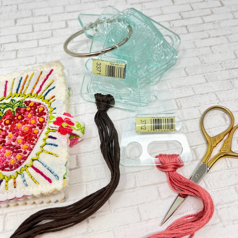 May include: Two clear plastic thread organizers with brown and pink embroidery floss. The organizers have labels with the numbers "3371" and "3712" and barcodes. A pair of gold scissors is also pictured.