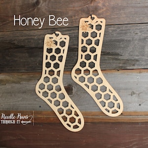 May include: Two wooden sock blockers shaped like a honeycomb with a bee design. The text "Honey Bee" is above the blockers. The text "A Needle Runs Through It" is below the blockers.