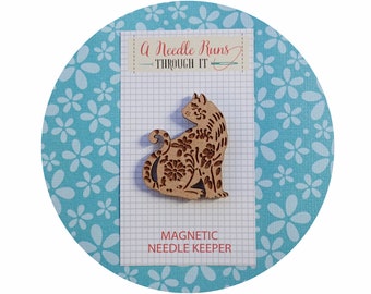 May include: A wooden cat-shaped magnetic needle keeper with a floral design. The text "A Needle Runs Through It" and "Magnetic Needle Keeper" are printed on the card behind the cat.