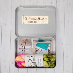 Może przedstawiać: Otwarta, srebrna puszka zawierająca materiały do robienia na drutach. W środku znajdują się drewniane narzędzia, turkusowy kawałek materiału, nożyczki i dwie motki przędzy w kolorze różowym, żółtym i zielonym. Na pokrywie znajduje się napis "A Needle Runs Through It".