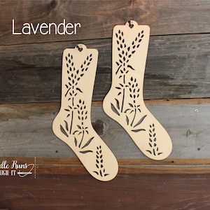 May include: Two wooden sock-shaped ornaments with a cut-out design of lavender flowers. The ornaments are hanging on a wooden wall. The text "Lavender" is written above the ornaments. The text "A Needle Runs Through It" is written below the ornaments.