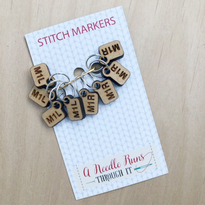 May include: A set of wooden stitch markers on a white card with the text "STITCH MARKERS" at the top. Each marker is a small, rectangular tag with "M1L" or "M1R" engraved on it. The card also has the text "A Needle Runs THROUGH IT".