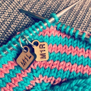 May include: Close-up of knitting needles with a turquoise and pink knitted fabric. Two small wooden tags with the letters "M1L" and "M1R" hang from the knitting. The fabric has a striped pattern.
