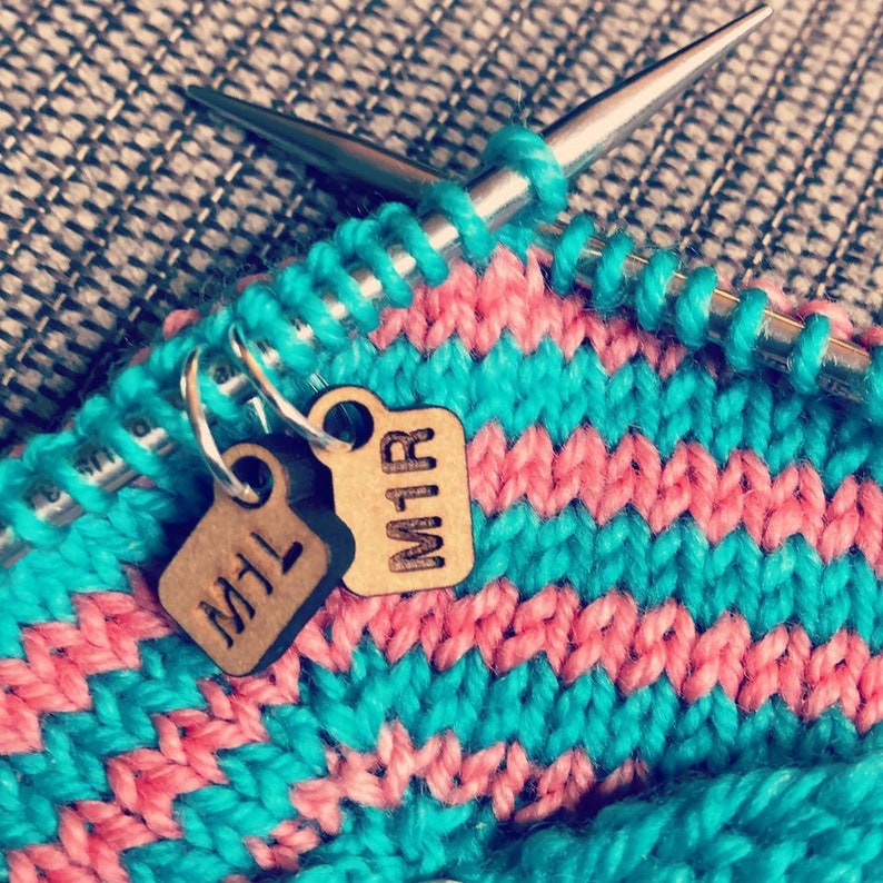 May include: Close-up of knitting needles with a turquoise and pink knitted fabric. Two small wooden tags with the letters "M1L" and "M1R" hang from the knitting. The fabric has a striped pattern.
