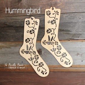 May include: Two wooden sock blockers featuring a hummingbird and floral design. The blockers are made of light brown wood and have a cutout design. The text "Hummingbird" is written at the top of the image.