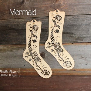 May include: Two wooden sock-shaped stencils featuring a mermaid design. The stencils are cut out with a mermaid, fish, seashells, and stars. The text "Mermaid" is at the top of the image.