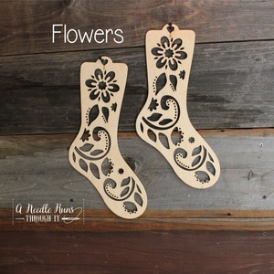 Puede incluir: Dos adornos de madera con forma de calcetín con recortes florales. Los adornos son de color marrón claro y presentan una flor en la parte superior y diseños intrincados de hojas y espirales. La palabra "Flowers" está escrita encima de los adornos.