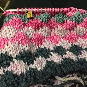 May include: Close-up of a knitting project in progress, featuring a vibrant color palette. The knitted fabric displays a textured pattern with alternating bands of pink, gray, and white yarn. A small wooden tag with the letters "BOR" hangs from the knitting needle.