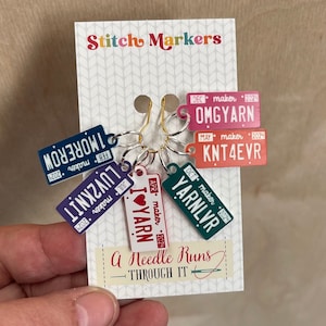 Könnte beinhalten: Sechs bunte Stichmarkierungen in Form von Nummernschildern mit verschiedenen Monaten und Herstellernamen. Die Markierungen lauten "OMGYARN", "KNT4EVR", "YARNLVR", "I LOVE YARN", "LUV2KNIT" und "1MOREROW". Der Text "A Needle Runs Through It" ist auf der Karte gedruckt.