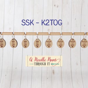 May include: Wooden knitting stitch markers labeled "SSK LEFT" and "K2TOG RIGHT" are suspended from a wooden bar. The text "SSK - K2TOG" is above the markers. A banner below reads "A Needle Runs THROUGH IT".