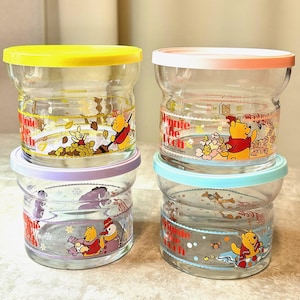 Puede incluir: Cuatro tarros de vidrio transparente con tapas de colores e ilustraciones de los personajes de Winnie the Pooh. Los tarros tienen tapas amarillas, rosas, moradas y azules. Cada tarro presenta una escena diferente con Pooh y sus amigos.