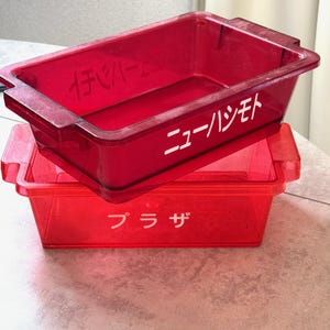 以下が含まれることがあります： 2つの透明な赤い長方形の収納容器が積み重ねられています。上の容器には白い文字で「ニューハシモト」と書かれています。下の容器には白い文字で「プラザ」と書かれています。両方の容器には、両側にハンドルが付いています。