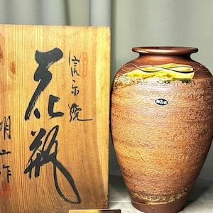 以下が含まれることがあります： テクスチャ加工された表面と広い開口部を持つ茶色のセラミック花瓶。花瓶の上部には黄色と緑の装飾バンドがあります。花瓶の左側には、日本の書道が施された木箱があります。
