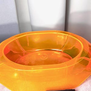 Op de afbeelding: Een doorschijnende, feloranje ovale plastic kom met een brede rand en gegoten structurele ribbels aan de buitenkant. De binnenkant van de bodem is voorzien van reliëftekst die deels zichtbaar is door het levendige, semi-transparante materiaal.