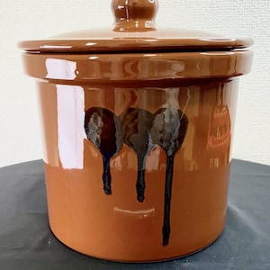 May include: A cylindrical brown ceramic crock with a matching lid and a glossy finish. The exterior features a decorative design of three dark brown, vertical drip-like patterns that extend downward from the upper section of the vessel.