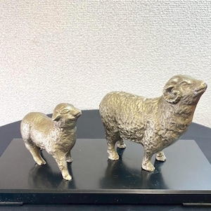 Puede incluir: Dos figuras de ovejas de metal en tono dorado sobre una base rectangular negra. La oveja más grande presenta cuernos curvados y un pelaje texturizado, mientras que la más pequeña está a su lado, ambas orientadas hacia la derecha.